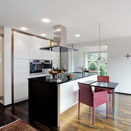 Eine moderne Küche mit weißen Schränken und schwarzen Arbeitsflächen. Eine Glasinsel mit einem Waschbecken und Wasserhahn sowie ein Esstisch mit roten Stühlen. Eine Lampe hängt von der Decke und ein Kreuz ist an der Wand.