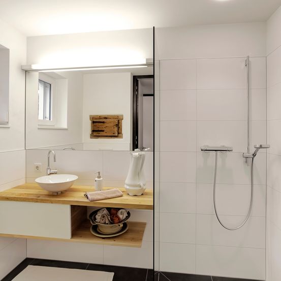 Ein modernes Badezimmer mit weißen Fliesen, einer Holzarbeitsplatte, einem runden Waschbecken und einer Duschkopf. Ein Spiegel reflektiert den Raum, und ein Holzkasten hängt an der Wand.