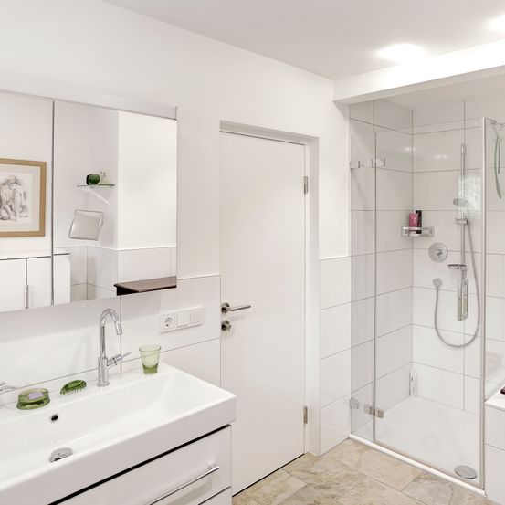 Ein modernes, komplett weißes Badezimmer mit Dusche, Waschbecken und großem Spiegel. Die Dusche hat eine Glastür und silberne Armaturen. Das Waschbecken hat einen Wasserhahn und einen grünen Becher.