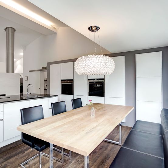 Eine moderne Küche mit Holztisch, schwarzen Stühlen und einer Kronleuchter. Weiße Schränke, eine Spüle und ein Ofen sind vorhanden. Der Boden ist aus Holz.