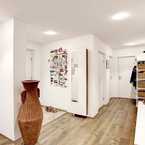 Ein Innenkorridor mit Holzboden und weißen Wänden. Eine große braune Vase steht links. Fotos und Gegenstände schmücken die Wand rechts.