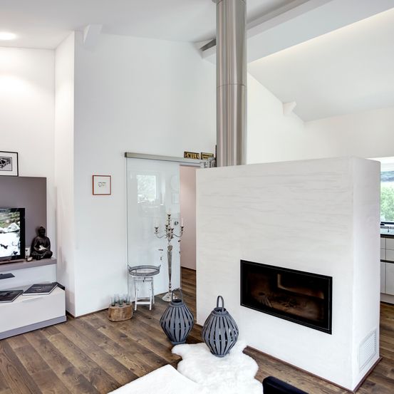 Innenraum eines modernen Wohnzimmers mit weißer Wand, Holzboden und Kamin. Ein Fernseher, Bücher und eine Buddha-Statue sind auf der linken Seite.