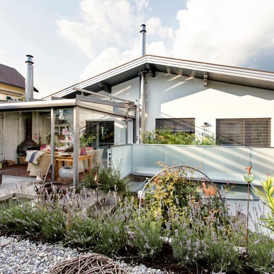 Ein weisses Haus mit einer Glaserweiterung, einem Balkon und einem Garten mit Lavendel und anderen Pflanzen.