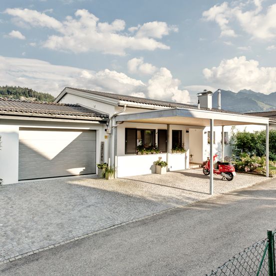 Ein modernes weißes Haus mit einem roten Roller, der unter einem Carport geparkt ist, in einer ruhigen Gegend mit Bergen im Hintergrund gelegen.