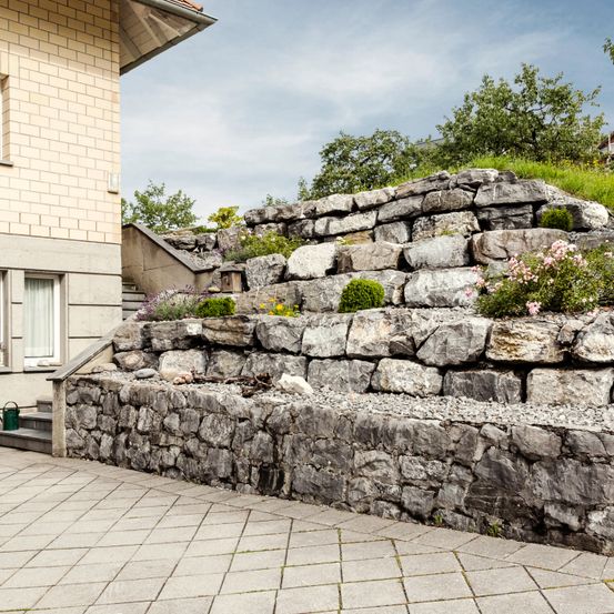 Ein Steinmauer mit Pflanzen und Blumen befindet sich neben einem Haus mit einer Treppe, die zum Eingang führt.