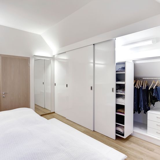 Ein minimalistisches Schlafzimmer mit weißen Wänden und Holzböden. Ein Bett steht zentral, und ein offener Kleiderschrank mit ordentlich organisierter Kleidung befindet sich rechts.