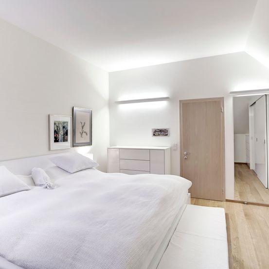 Ein minimalistisches Schlafzimmer mit weißen Wänden, einem Holzbett und einer Holztür. Über dem Bett hängen zwei gerahmte Bilder. Ein weißer Schrank mit Schubladen steht neben dem Bett.