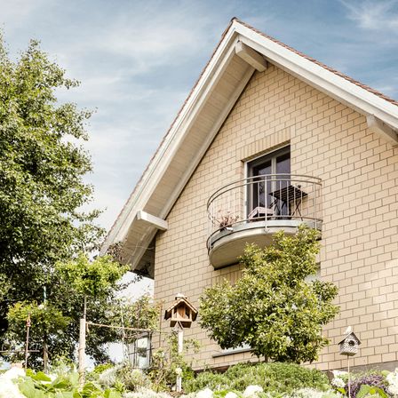 Ein Haus mit Balkon, Ziegelwänden und einem Ziegeldach. Ein Balkon hat einen Tisch und Stühle. Der Garten hat ein Vogelhaus, Bäume und Blumen.
