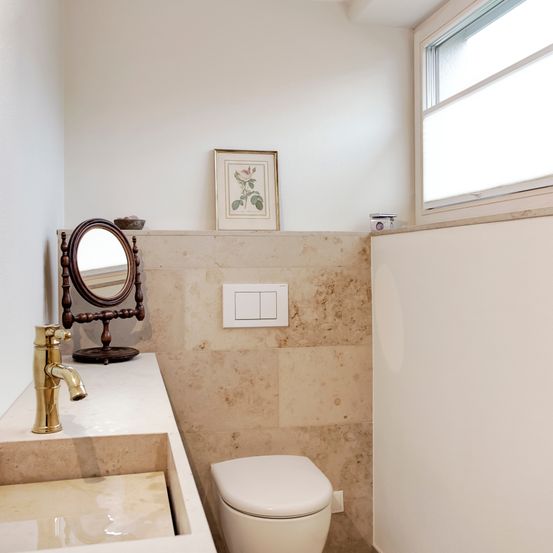 Ein Badezimmer mit einem weißen Toilettensitz, einem Waschtisch mit einem goldenen Wasserhahn, einem Wandspiegel und einem gerahmten Bild über der Toilette. Die Wand ist beige mit Fliesen.