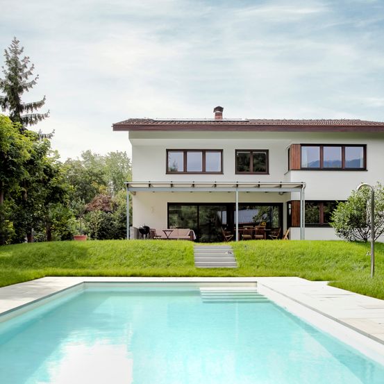Ein modernes weißes Haus mit einem Ziegeldach und einem großen Swimmingpool davor. Das Haus hat eine Glaswand mit Holzrahmen.