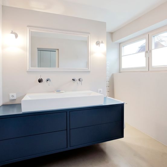 Ein Badezimmer mit einem weißen Waschbecken, einem Spiegel und einem blauen Schrank mit Schubladen. Es gibt zwei an der Wand montierte Lampen über dem Spiegel und eine Steckdose unter dem Waschbecken. Die Wand und die Decke sind weiß.