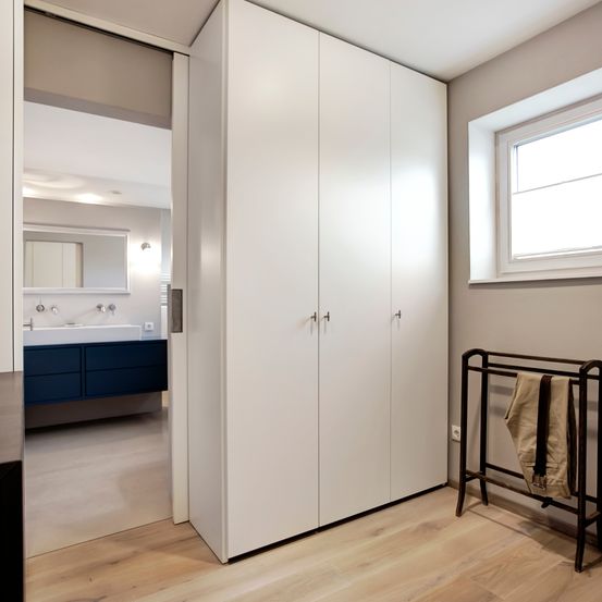 Modernes Badezimmer mit weißen Schränken, einem blauen Waschbecken, einem Spiegel und einem Fenster. Der Boden ist aus Holz und ein Handtuch liegt auf einem Metallständer.