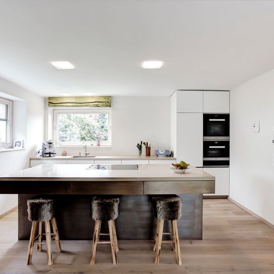 Eine moderne Küche mit einer großen Insel, Stühlen mit Fell und weißen Schränken. Ein Fenster mit Blick auf Bäume und eine Uhr an der Wand.