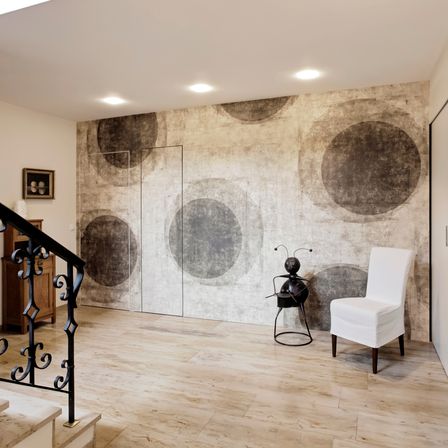 Ein moderner Raum mit einer großen Wand, die kreisförmige Muster aufweist, und einem Holzstuhl. Eine kleine Skulptur und ein gerahmtes Bild schmücken den Raum. Der Raum hat Holzböden und ein schwarzes Geländer.