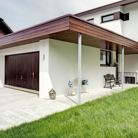 Ein modernes Haus mit einer braunen Garagentür und einem Carport, mit einer Bank, einem Tisch und einer Lampe auf dem Rasen.