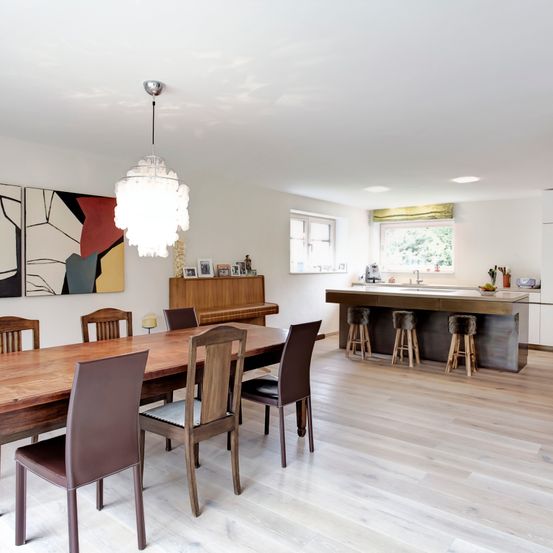 Ein offenes Wohnbereich mit Esstisch, Stühlen und moderner Pendelleuchte. Der Raum verfügt über Holzböden und weiße Wände. Ein Klavier und Kunstwerke im Hintergrund.