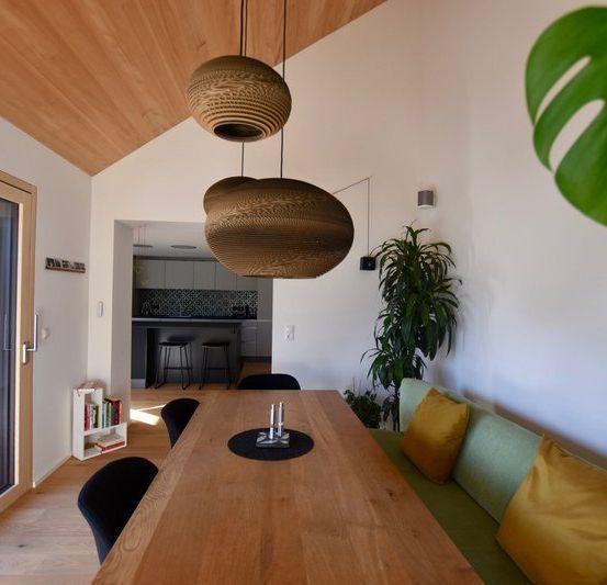 Ein gemütlicher Essbereich mit einem Holztisch, zwei Lampen und einer grünen Couch. Eine Topfpflanze steht rechts.