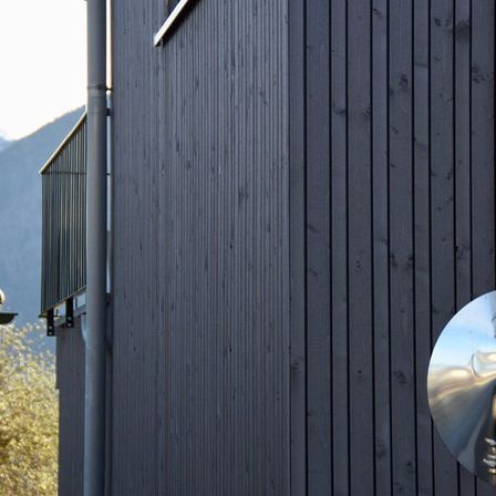 Ein modernes Haus mit schwarzer Holzverkleidung, einer Metallbrüstung und einem Metallrohr vor einem Berghintergrund.