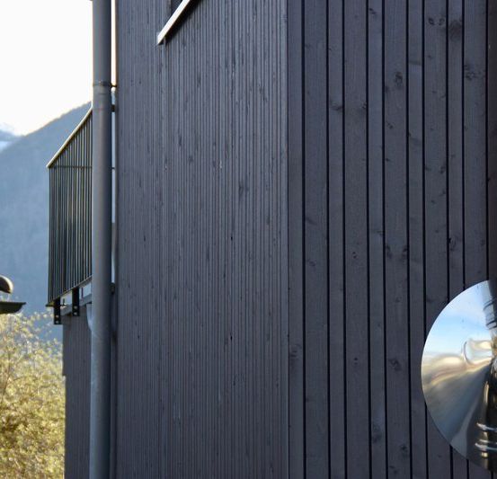 Ein modernes Haus mit schwarzer Holzverkleidung, einer Metallbrüstung und einem Metallrohr vor einem Berghintergrund.