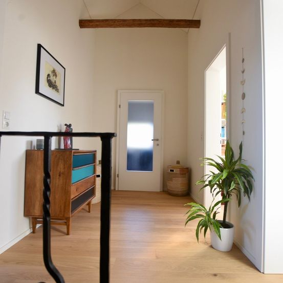 Ein leerer Flur mit einem Holzschrank, einem Bilderrahmen und einer Topfpflanze auf einem Holzboden. Die Wand ist weiß gestrichen, und eine Holzbalkendecke ist an der Decke montiert.