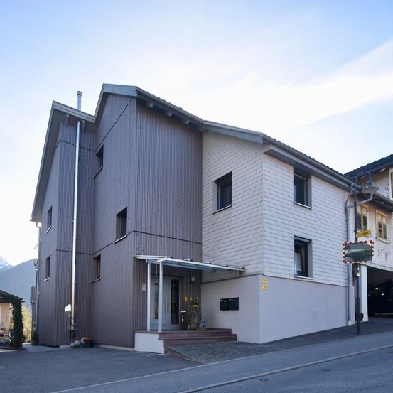 Ein modernes Mehrfamilienhaus mit geneigtem Dach und einer Mischung aus grauen und weißen Außenfassaden. Es hat mehrere Fenster und ein Glasvordach über dem Eingang. Es gibt Topfpflanzen auf den Stufen.