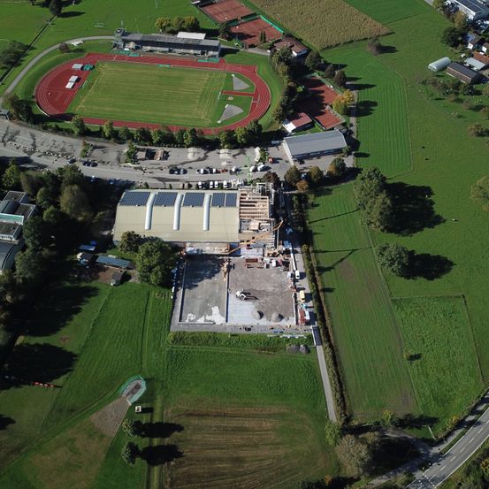 Luftaufnahme einer großen Baustelle mit grünen Feldern, einer roten Laufbahn und mehreren Gebäuden. Bäume und Autos sind verstreut.