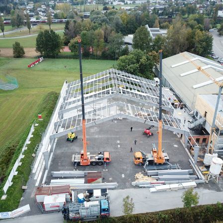 Luftaufnahme einer Baustelle mit Kränen, Arbeitern und Materialien. Die Baustelle zeigt ein Gebäude im Bau mit einem Metalldach. Verschiedene Maschinen und Vorräte sind auf der Baustelle positioniert.