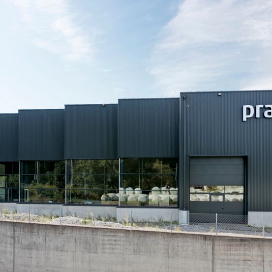 Ein modernes Industriegebäude mit grauen Wänden und reflektierenden Fenstern. Die Vorderseite ist mit einer großen Eingangstür und dem Wort 'pra' auf der Wand gekennzeichnet.