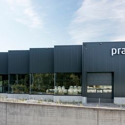 Ein modernes Industriegebäude mit grauen Wänden und reflektierenden Fenstern. Die Vorderseite ist mit einer großen Eingangstür und dem Wort 'pra' auf der Wand gekennzeichnet.