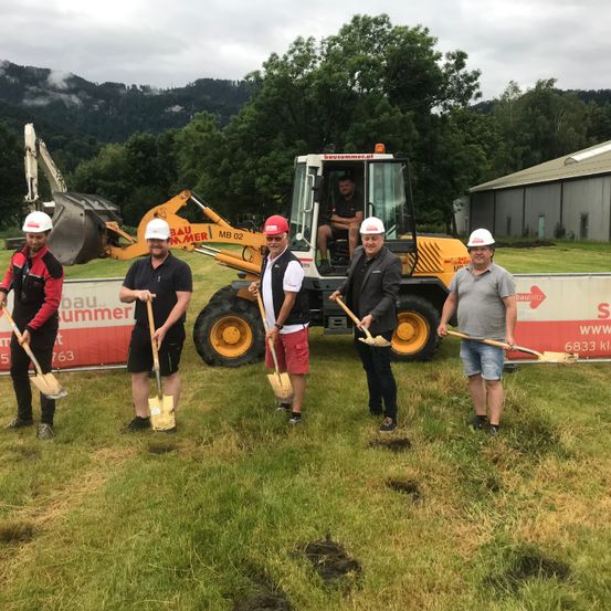 Fünf Männer stehen mit Schaufeln auf einem Feld, einer in einem grauen Shirt, einer in einem roten Shirt, zwei in Schwarz und einer in einem roten Hut. Ein Baufahrzeug ist im Hintergrund.