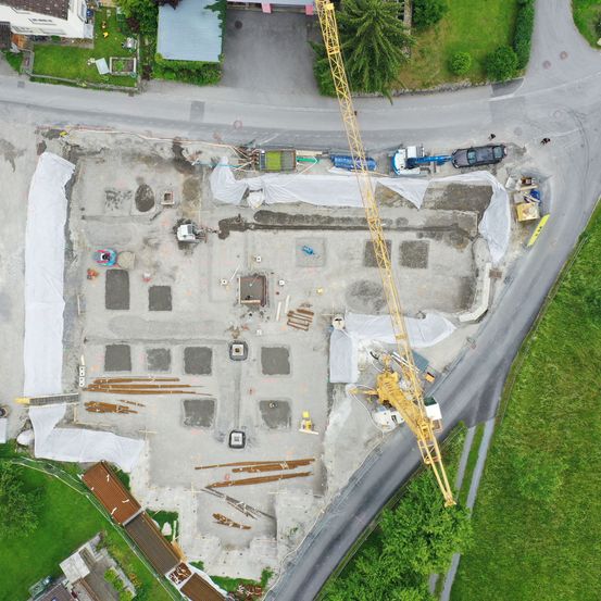 Eine Draufsicht auf eine Baustelle mit Kränen, Fahrzeugen und abgedeckten Materialien, umgeben von Grünflächen und einer Straße.