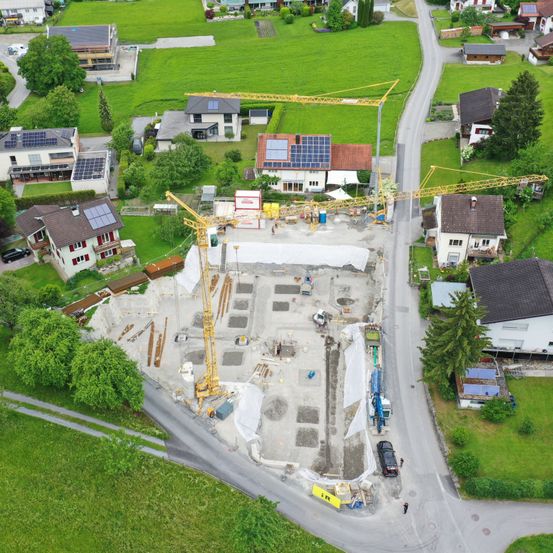 Luftaufnahme einer Wohnbaustelle mit Kränen, Baumaterialien und mehreren Häusern in einem grünen Viertel.