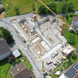 Eine Luftaufnahme einer Baustelle mit mehreren Kränen und Arbeitern, umgeben von Häusern, Straßen und Grünflächen.
