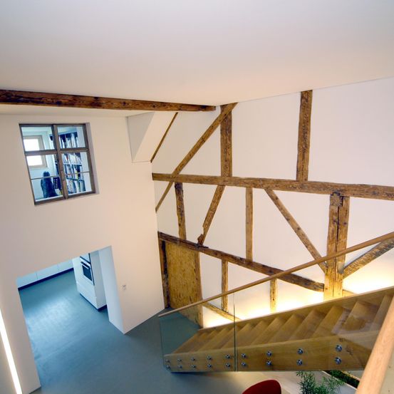 Ein Innenansicht eines Hauses mit Holzbalken und Glastreppe. Ein Fenster mit einer Person in der Nähe und einer weißen Wand.