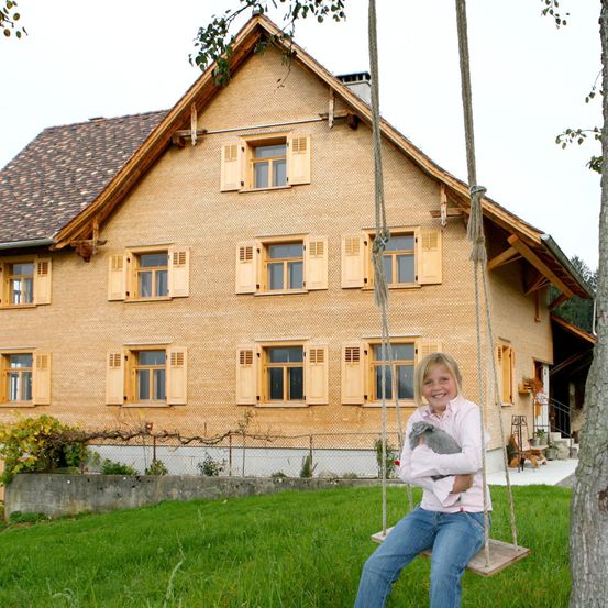 Ein junges Mädchen mit blonden Haaren sitzt auf einer Schaukel und hält einen grauen Hasen, vor einem Haus mit Holzwänden und gelben Fensterläden.