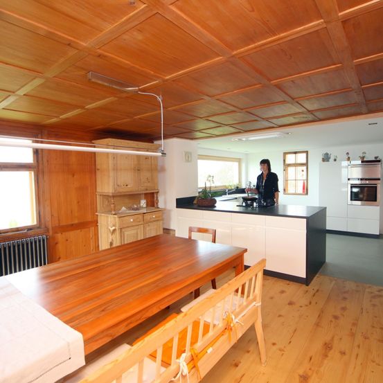 Eine Frau steht in einer Küche mit Holzböden und Schränken. Ein großer Holzesstisch steht vor einer Theke.