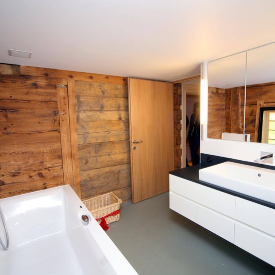 Ein Badezimmer mit Holzwänden und einer großen Badewanne, einem weißen Waschbecken und einem Spiegel. Ein Korb steht neben der Badewanne. Eine Person ist im Spiegel zu sehen.