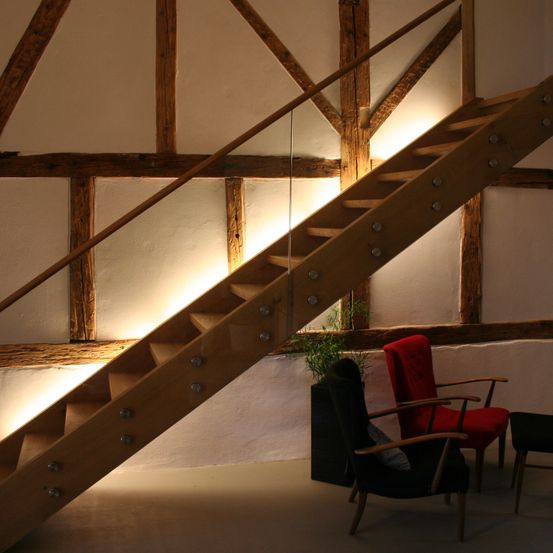 Eine Holztreppe mit einer Glasbrüstung, die zu einem Raum mit einer Holzdecke, Holzbalken und einem roten Stuhl führt. Ein Topf mit einer Pflanze steht daneben.