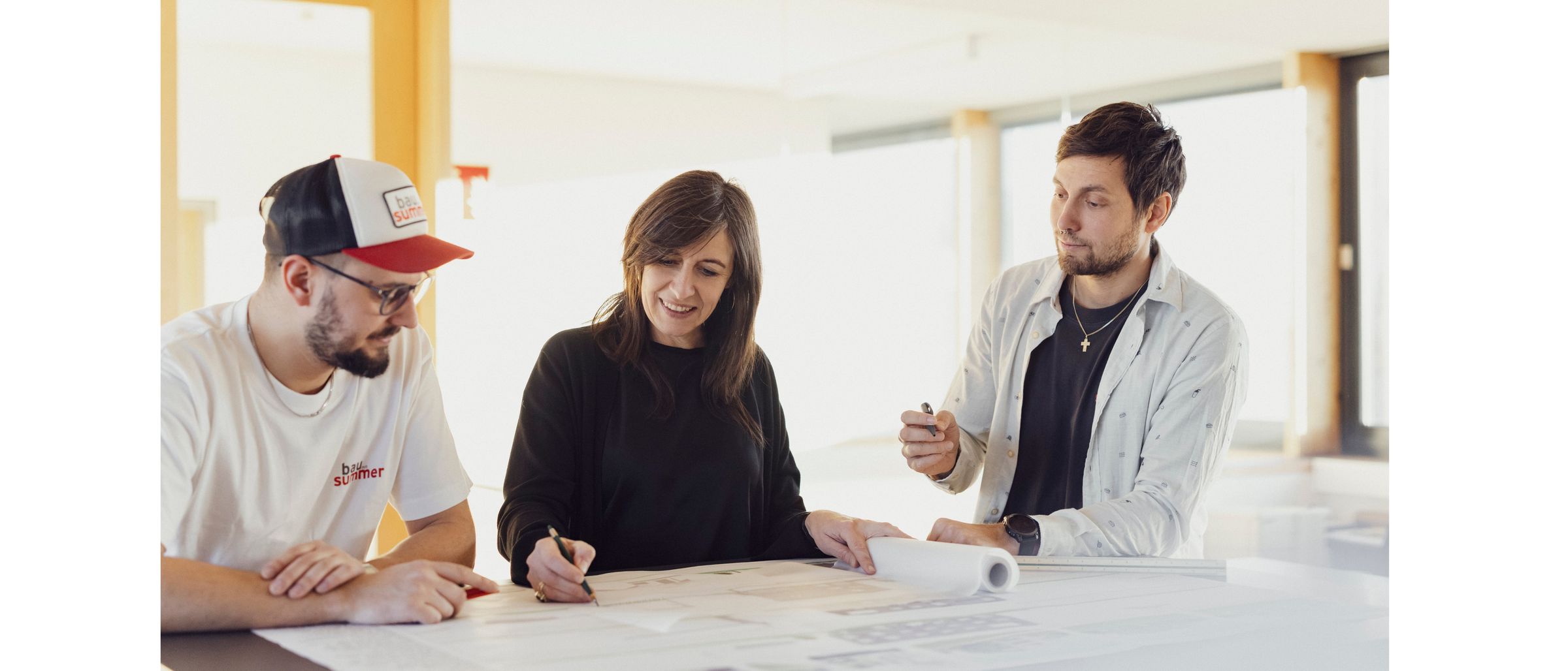 Drei Menschen arbeiten gemeinsam an Blaupausen, an einem Tisch sitzend, wobei eine Frau einen Stift hält.