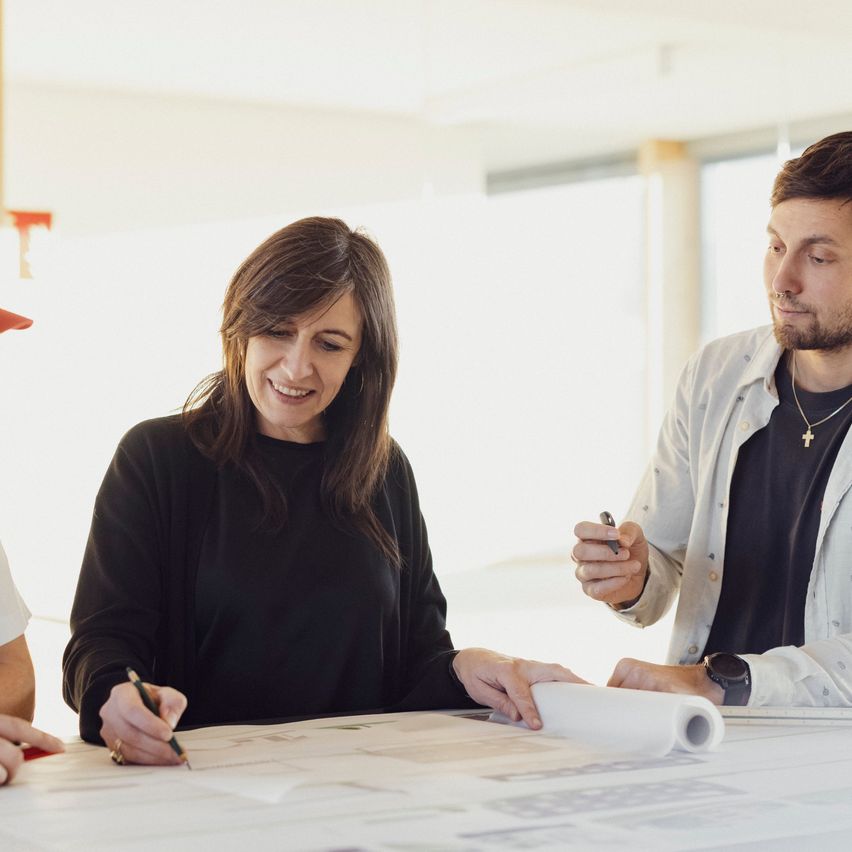 Drei Menschen arbeiten gemeinsam an Blaupausen, an einem Tisch sitzend, wobei eine Frau einen Stift hält.
