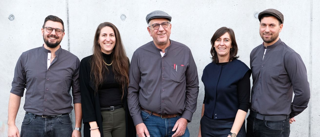 Fünf Personen stehen zusammen vor einer weißen Wand und lächeln für ein Foto. Der Mann in der Mitte trägt Hut und Brille.