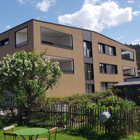 Ein modernes, mehrstöckiges Gebäude mit mehreren Balkonen, umgeben von einem Garten mit Zaun und blühenden Bäumen. Das Gebäude hat eine braune Außenseite und mehrere Fenster. Ein grüner Tisch und Stühle sind im Garten aufgestellt.