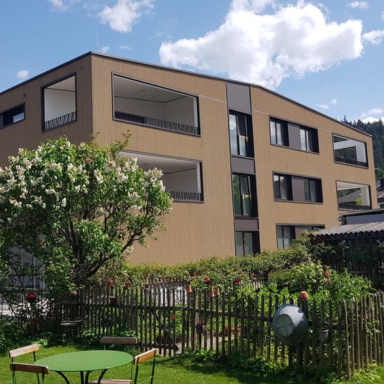 Ein modernes, mehrstöckiges Gebäude mit mehreren Balkonen, umgeben von einem Garten mit Zaun und blühenden Bäumen. Das Gebäude hat eine braune Außenseite und mehrere Fenster. Ein grüner Tisch und Stühle sind im Garten aufgestellt.