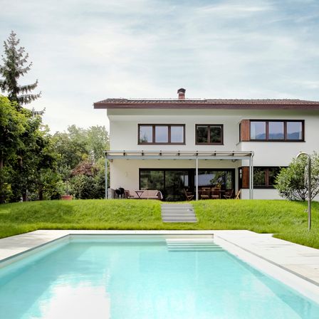 Ein modernes weißes Haus mit einem braunen Dach steht in einem üppigen grünen Garten. Das Haus hat große Fenster, eine Terrasse und einen Swimmingpool vor der Fassade. Bäume umgeben das Grundstück.