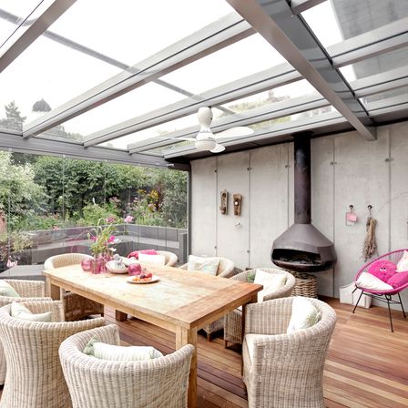 Ein moderner Essbereich im Freien mit Holzböden, Korbstühlen, einem Holztisch, einer Glaswand, einem Deckenventilator und einem Schornstein.