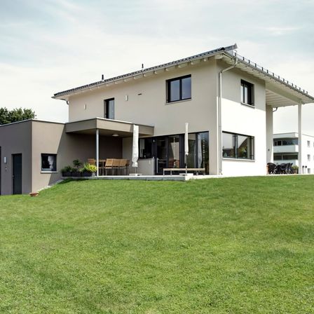 Ein modernes zweistöckiges Haus mit großem Rasen davor, ausgestattet mit großen Fenstern und einer Terrasse mit Tisch und Stühlen.