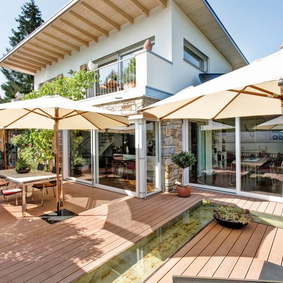 Ein modernes Haus mit einer Holzterrasse hat große Sonnenschirme, einen Esstisch mit Blumen und ein Wasserelement. Es hat Glaswände und -türen und einen Balkon mit Geländern.