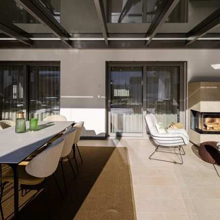 Ein elegantes Innen-Esszimmer mit einem langen Tisch und Stühlen, großen Glastüren und einem Kamin mit Holz. Der Raum hat ein modernes Design mit einer Betondecke und gefliestem Boden.