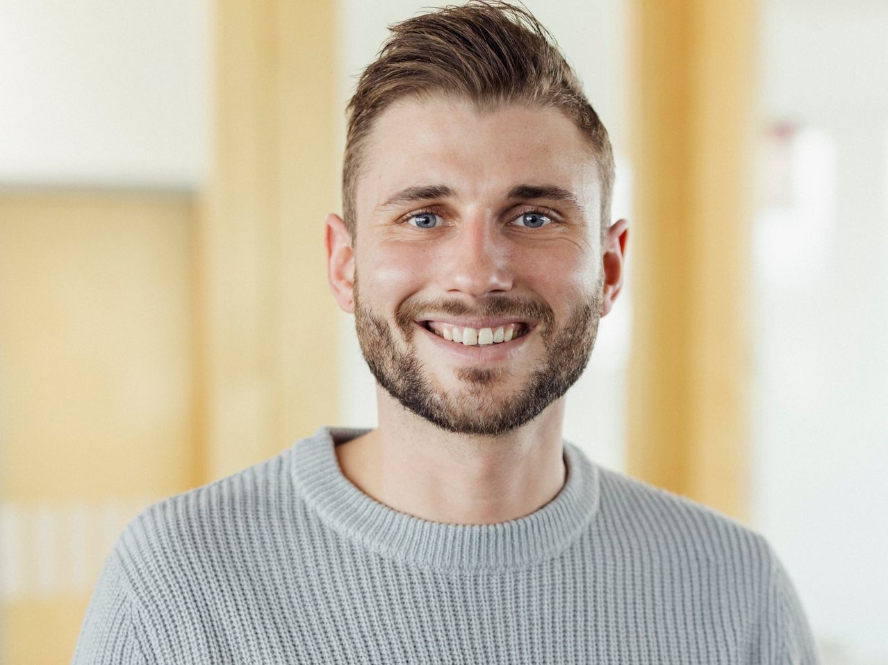 Ein Mann mit Bart und Oberlippenbart lächelt fröhlich, trägt einen grauen Pullover und hat blaue Augen.