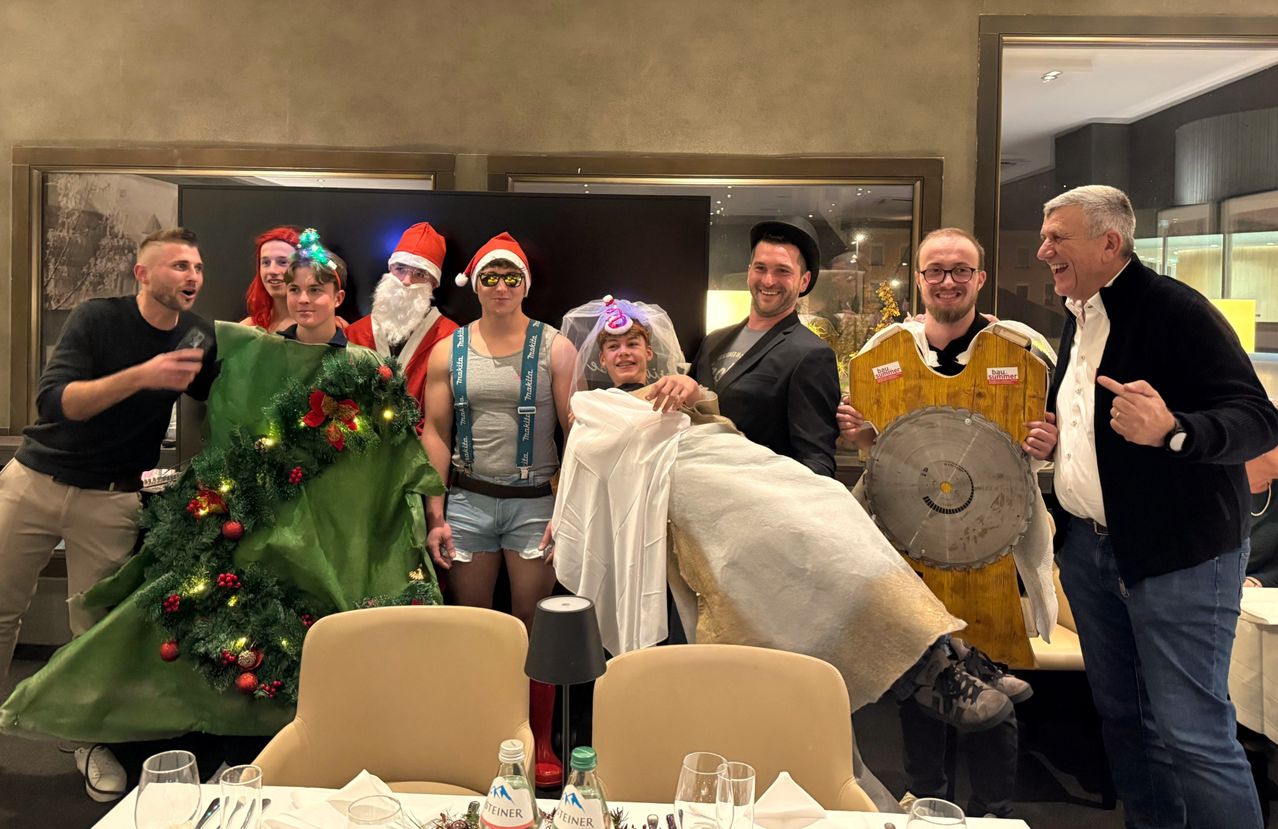 Eine Gruppe von Menschen in verschiedenen Kostümen, darunter der Weihnachtsmann, eine Braut und andere, stehen in einem Raum, lächeln und posieren für ein Foto. Dahinter steht ein geschmückter Weihnachtsbaum und ein Tisch, der für eine Mahlzeit gedeckt ist.